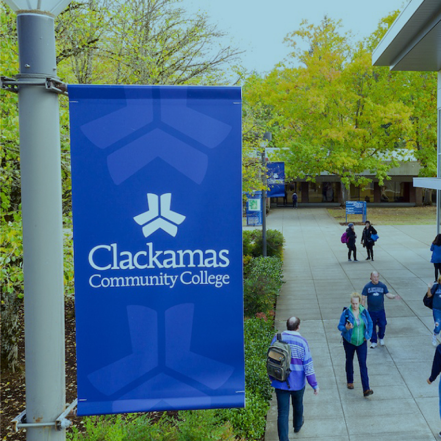 A school flag connected to a pole beside a walkway full of students going to and from school that says "Clackamas Community College"