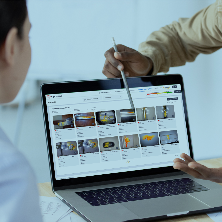 A man in a khaki long sleeve shirt is pointing at images from the Optisolve Reports dashboard with a pencil to another co-worker for training